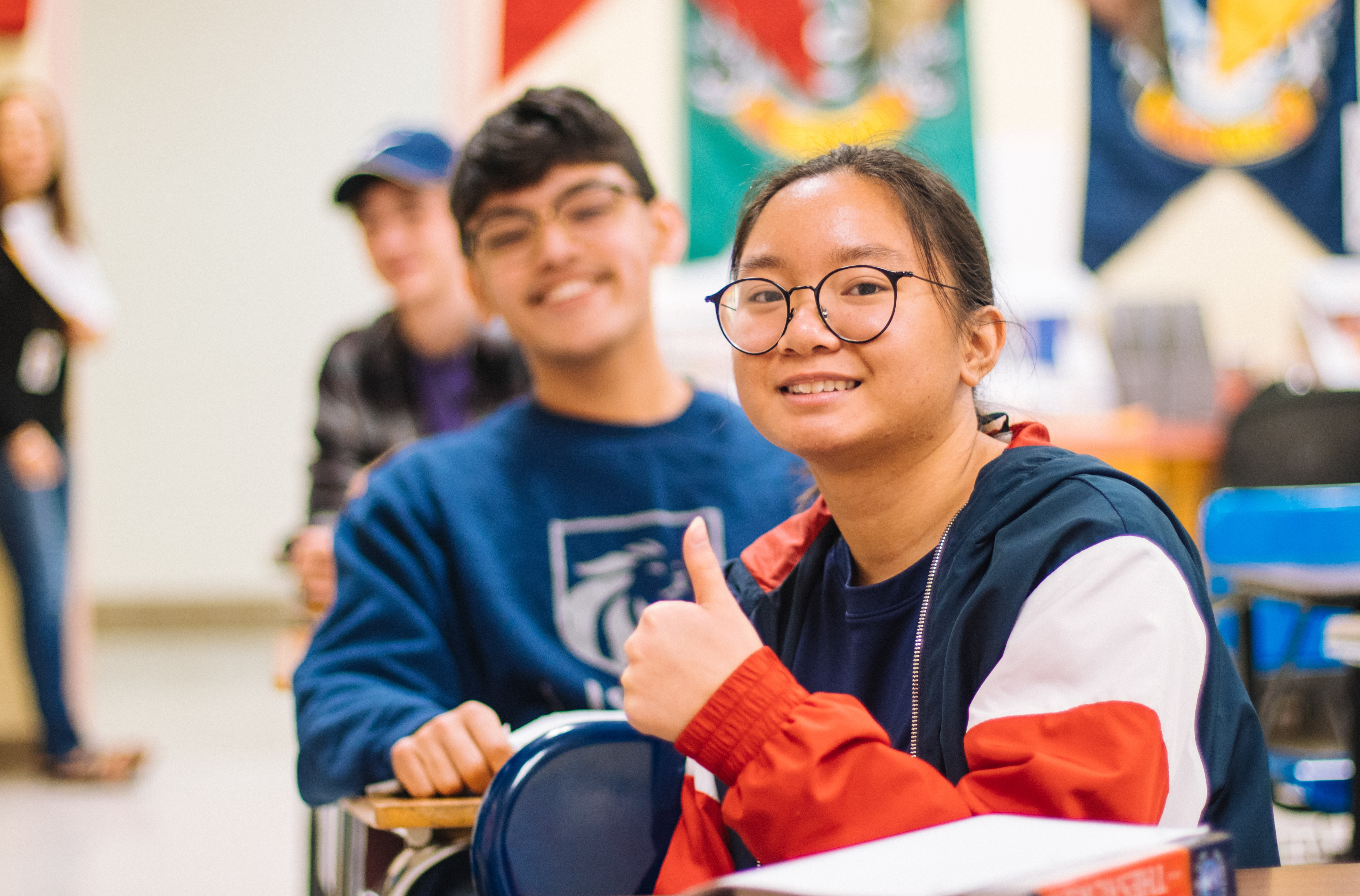 Two students inside of a classroom, smiling and giving a thumbs up